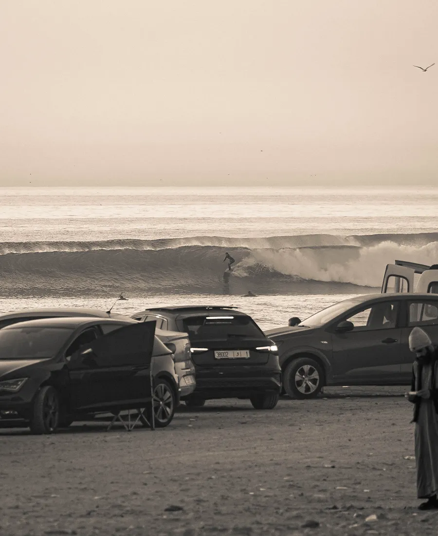 Surfers riding waves at Anza Beach, photographed from the coastal road above parked cars