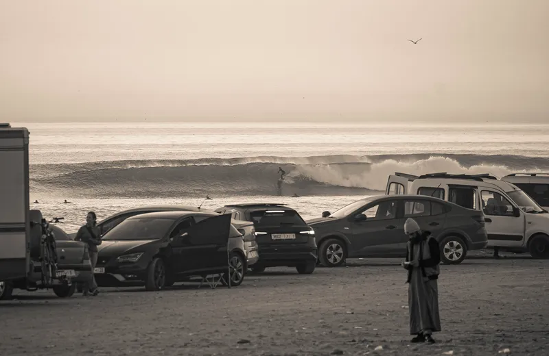 Surfers riding waves at Anza Beach, photographed from the coastal road above parked cars