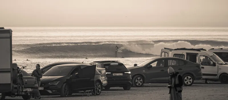 Surfers riding waves at Anza Beach, photographed from the coastal road above parked cars