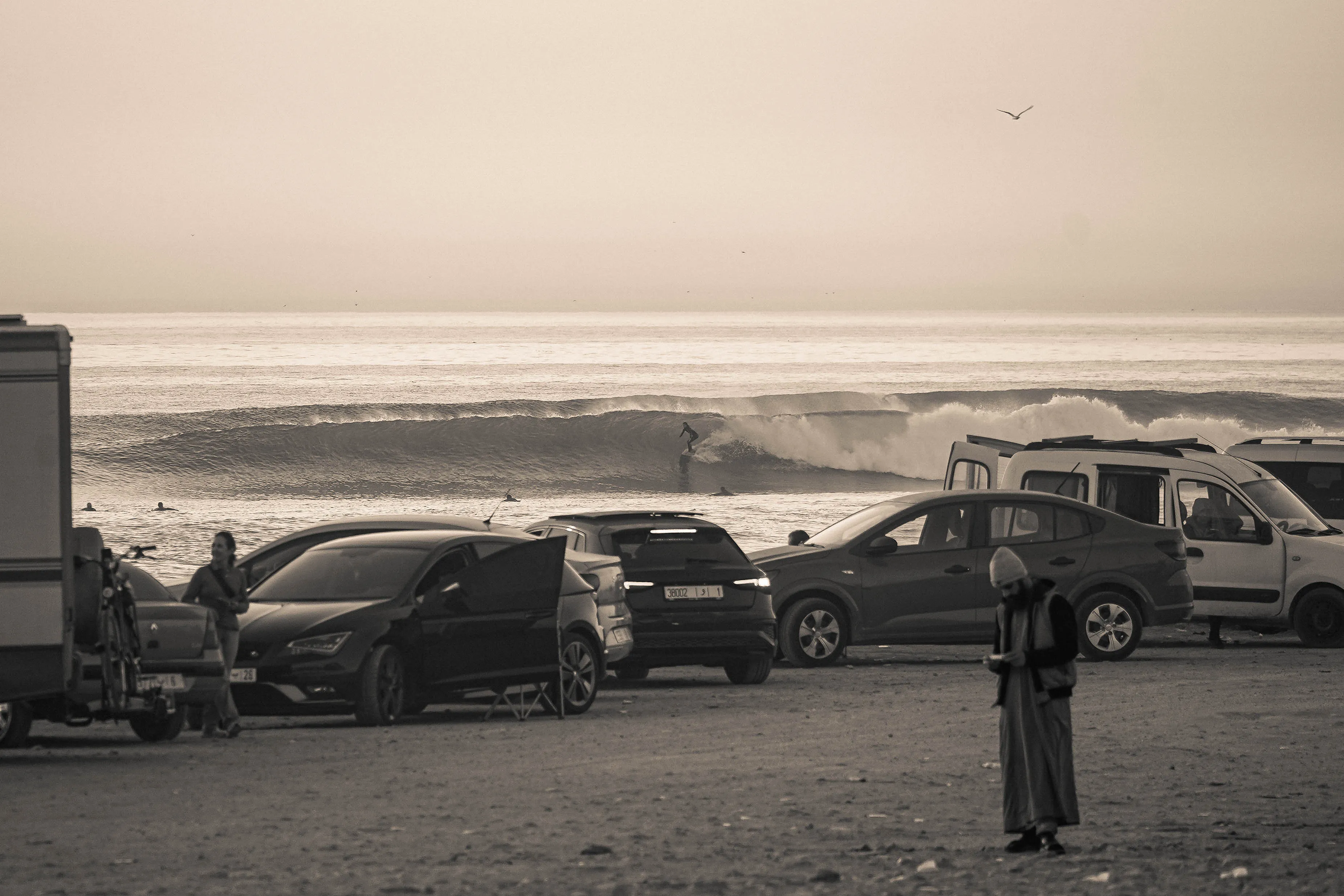Surfers riding waves at Anza Beach, photographed from the coastal road above parked cars