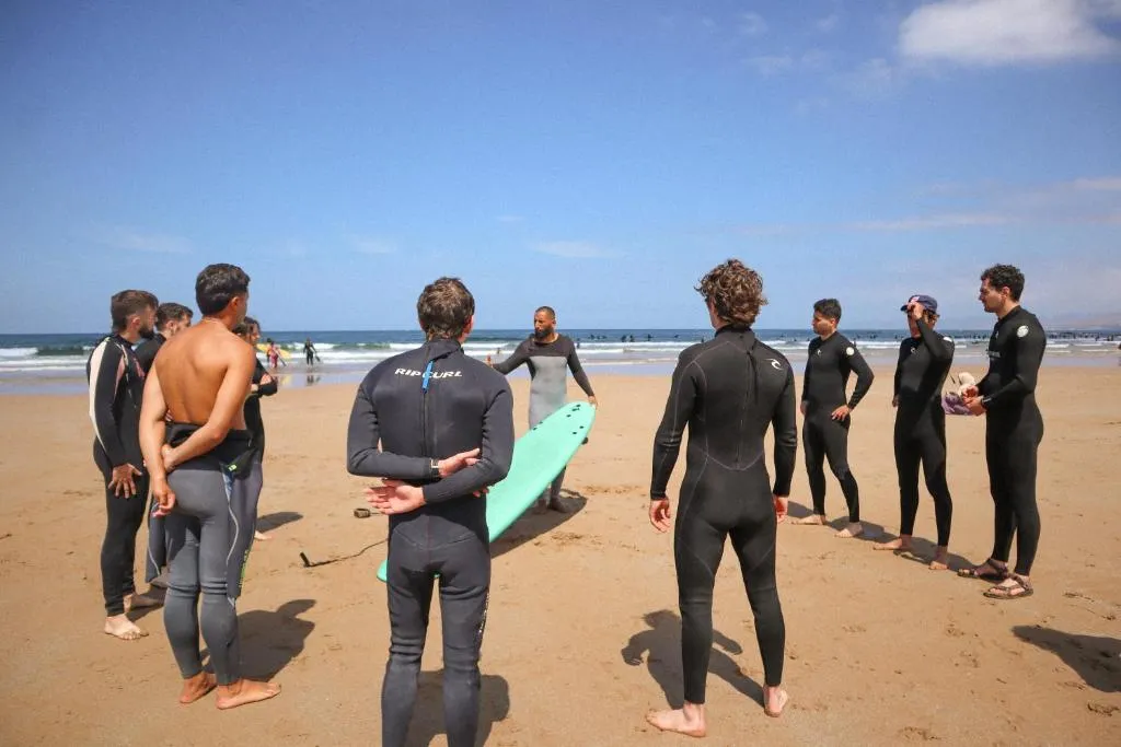 Jamal coaching a circle of surf-week guests in the sand, explaining tide and lineup positioning